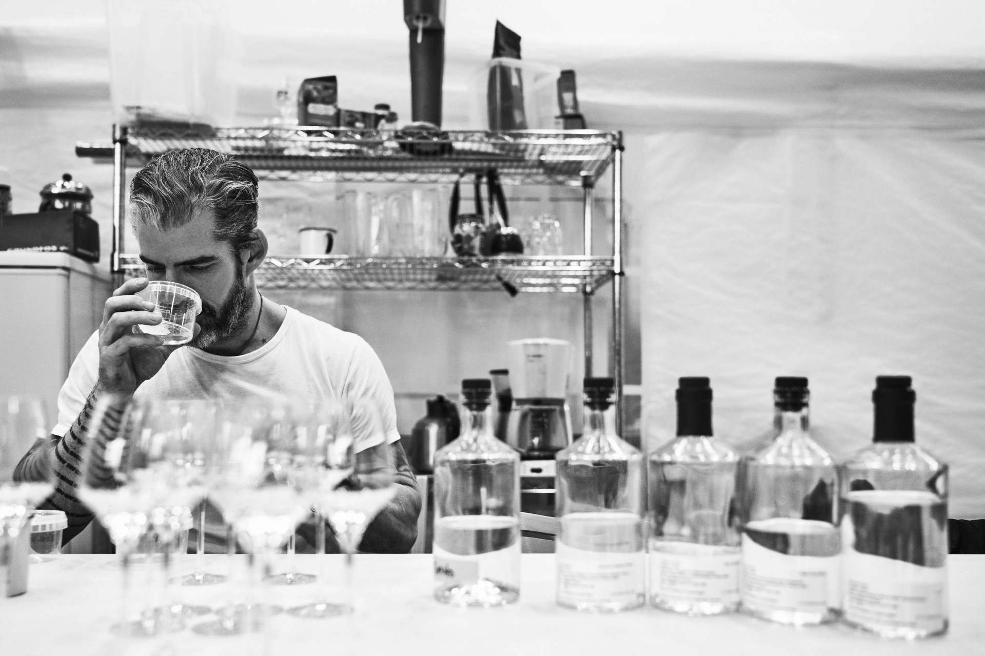 man sitting in front of a table full of bottles and glasses, tasting the contents of one of the glasses