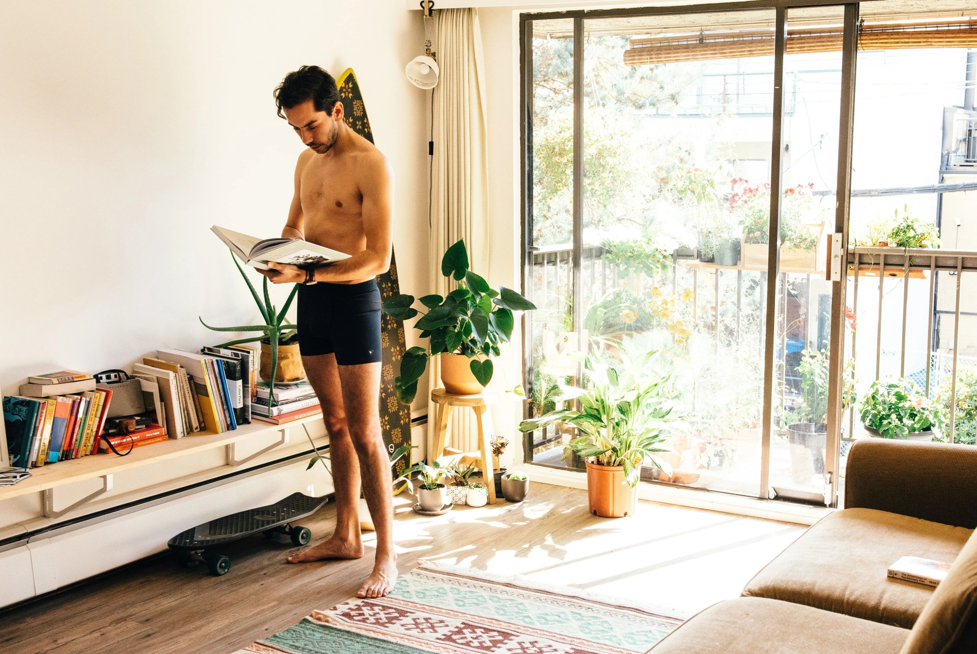 man standing in a sunny room, reading a book in front of a bookshelf