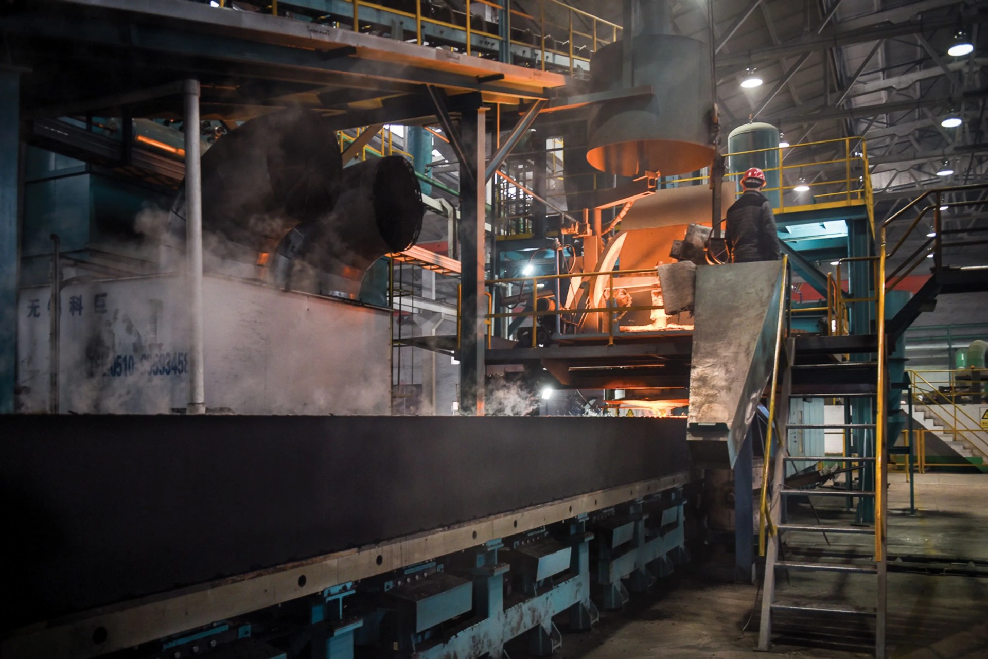 inside of a factory showing multiple metal machines, steam, and a man with a hardhat