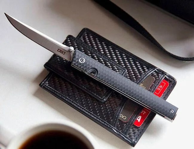 a slim folding pocket knife atop a black wallet on a table next to a cup of coffee