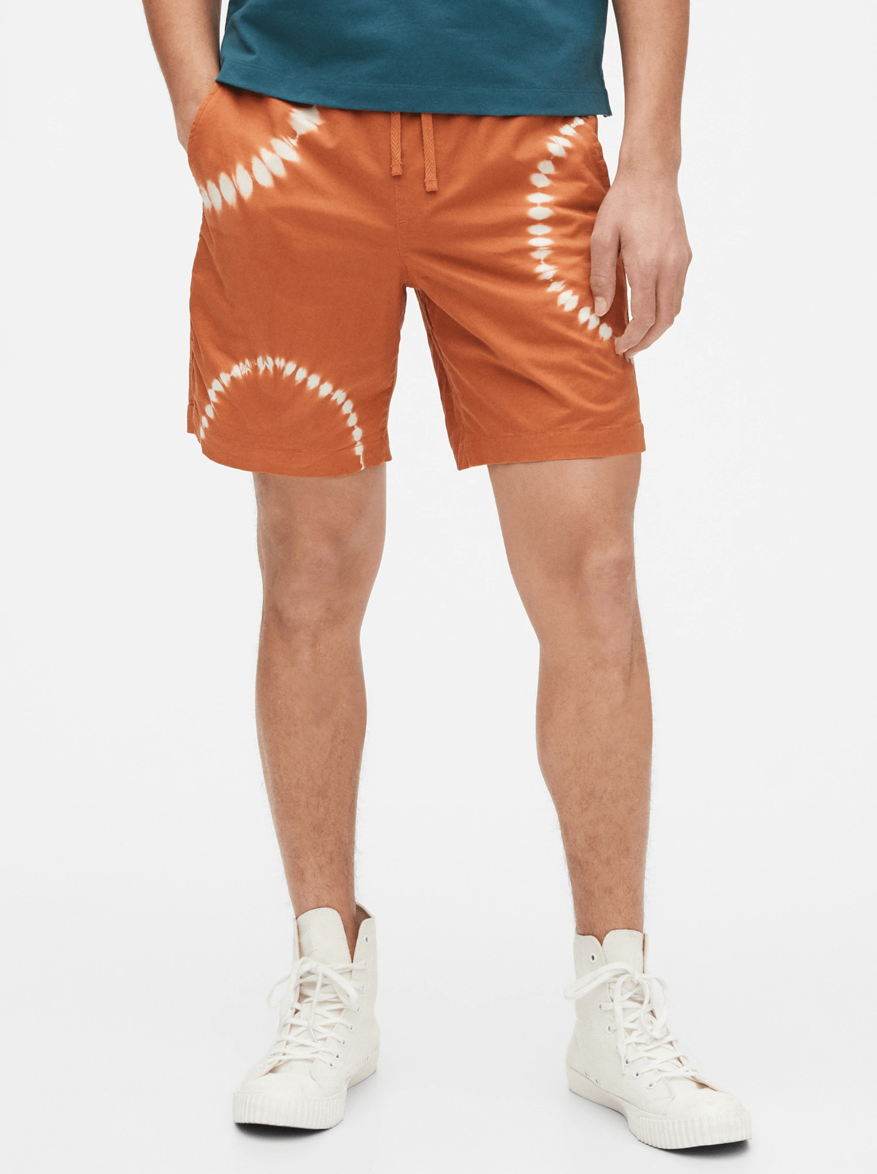 Orange tie dye shorts on a model with a blue shirt and white high-top sneakers on a white background.