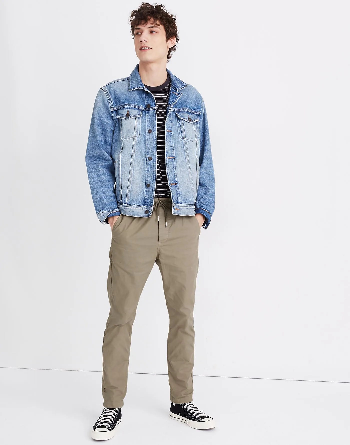 Olive twill pants on a model with a denim jacket, striped shirt, black and white sneakers on a gray background.