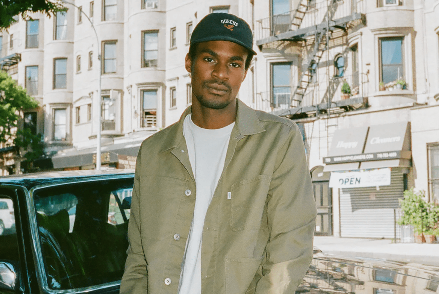 a model sitting on a car wearing a black hat, white t shirt, olive chore coat and blue jeans