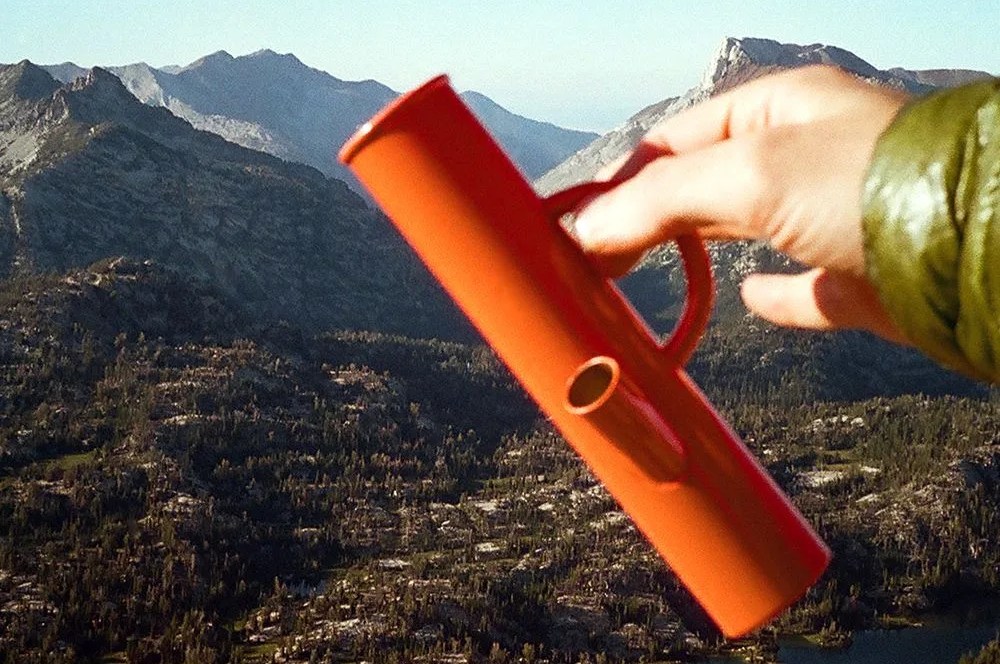 a hand holding an orange bong against a mountain backdrop