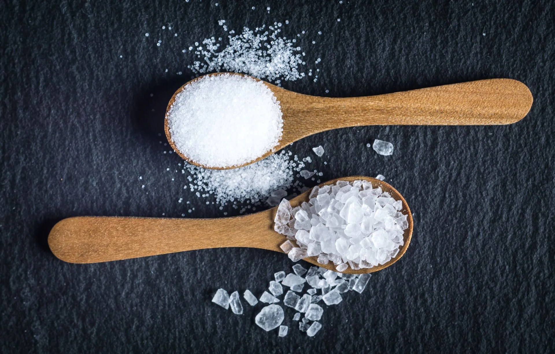 different types of salt sea and kitchen salt top view on two wooden spoons on black background