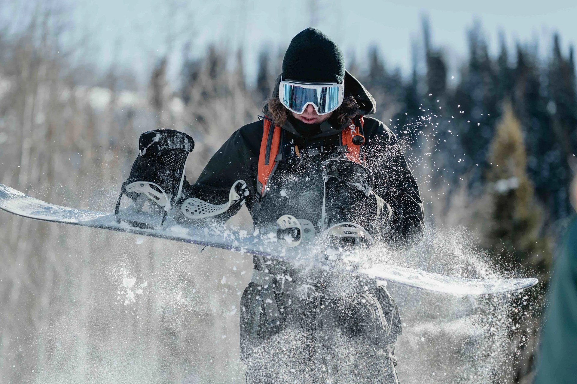 man wearing winter gear shaking snow off his snowboard