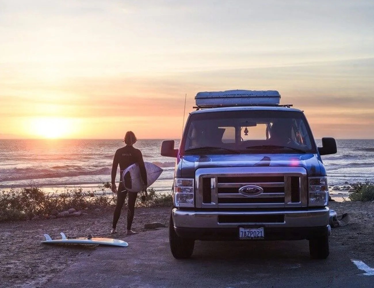 man at beach with surfboard next to an escape campervan