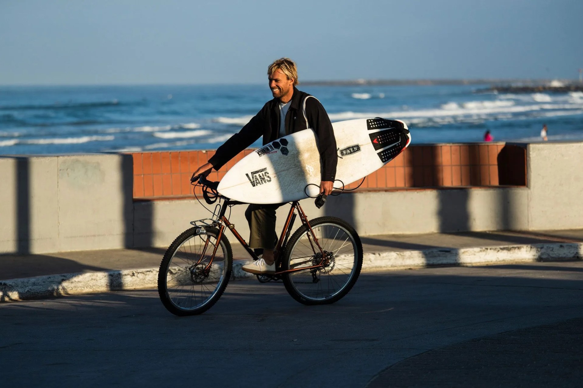 man wearing vans shoes riding bicycle holding surfboard by beach