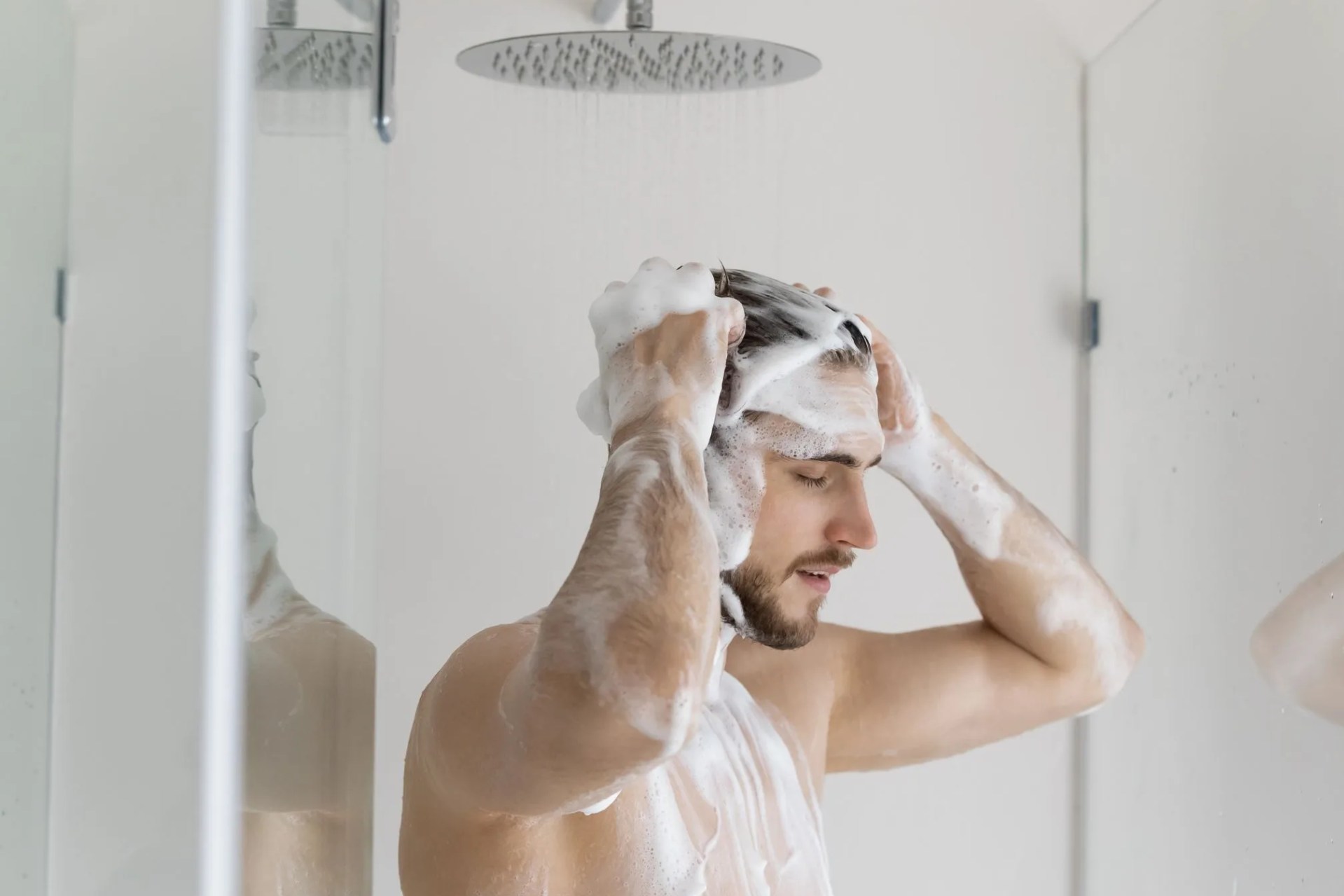 calm relaxed handsome young man with muscular torso and foamy head taking shower, washing hair with antidandruff shampoo, standing under showerhead with pouring water, caring for routine hygiene