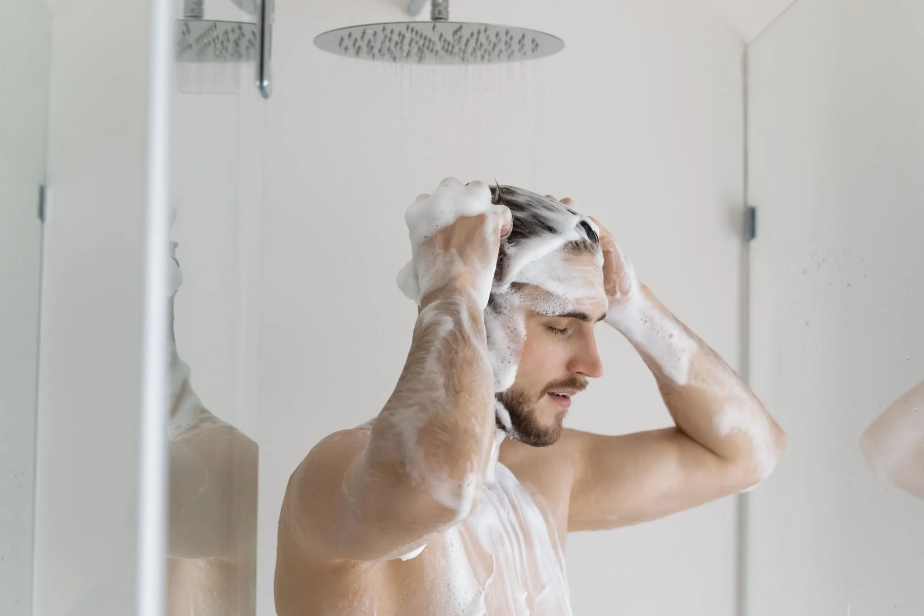 calm relaxed handsome young man with muscular torso and foamy head taking shower, washing hair with antidandruff shampoo, standing under showerhead with pouring water, caring for routine hygiene