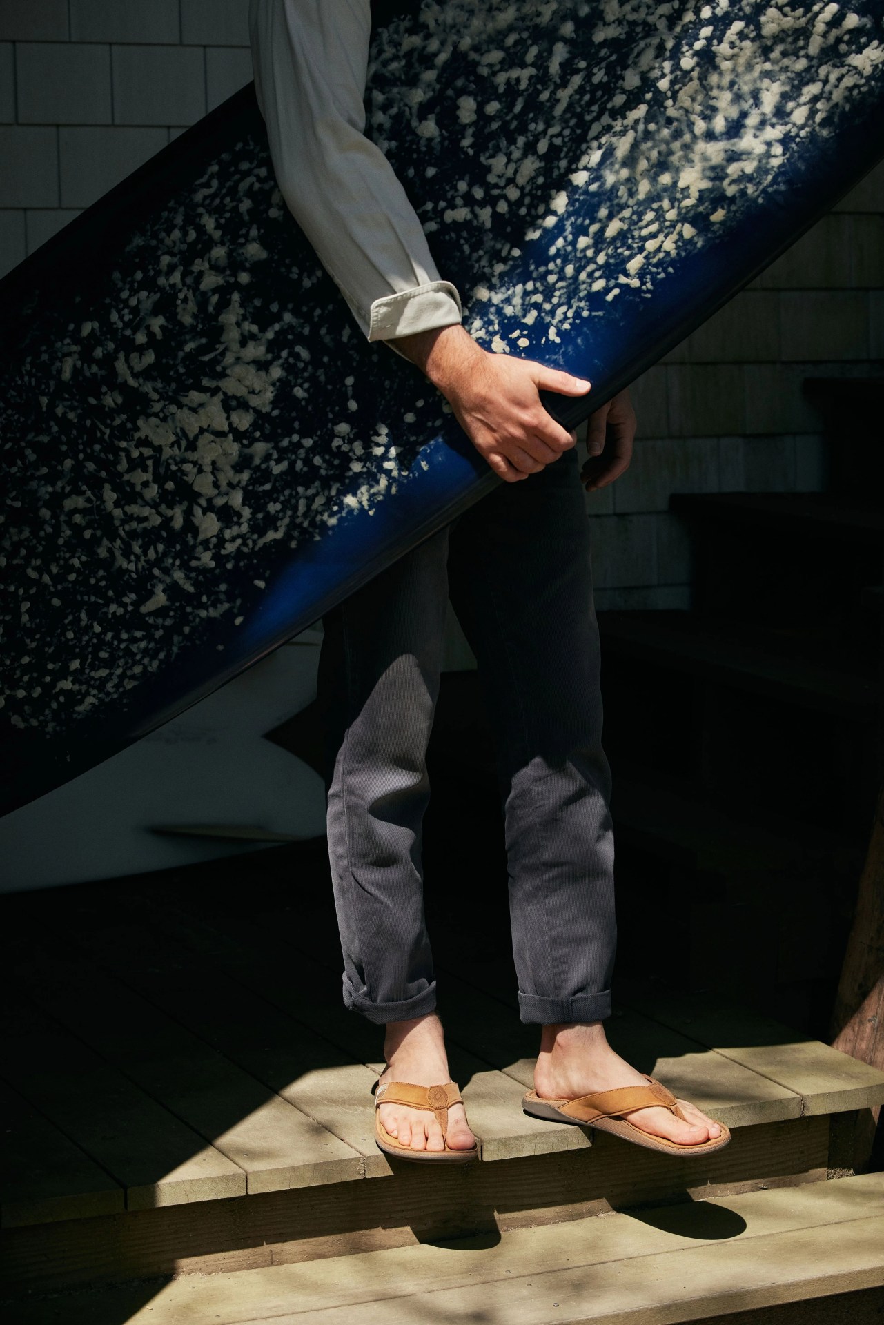 man holding surfboard with olukai tuahine sandals worn on feet