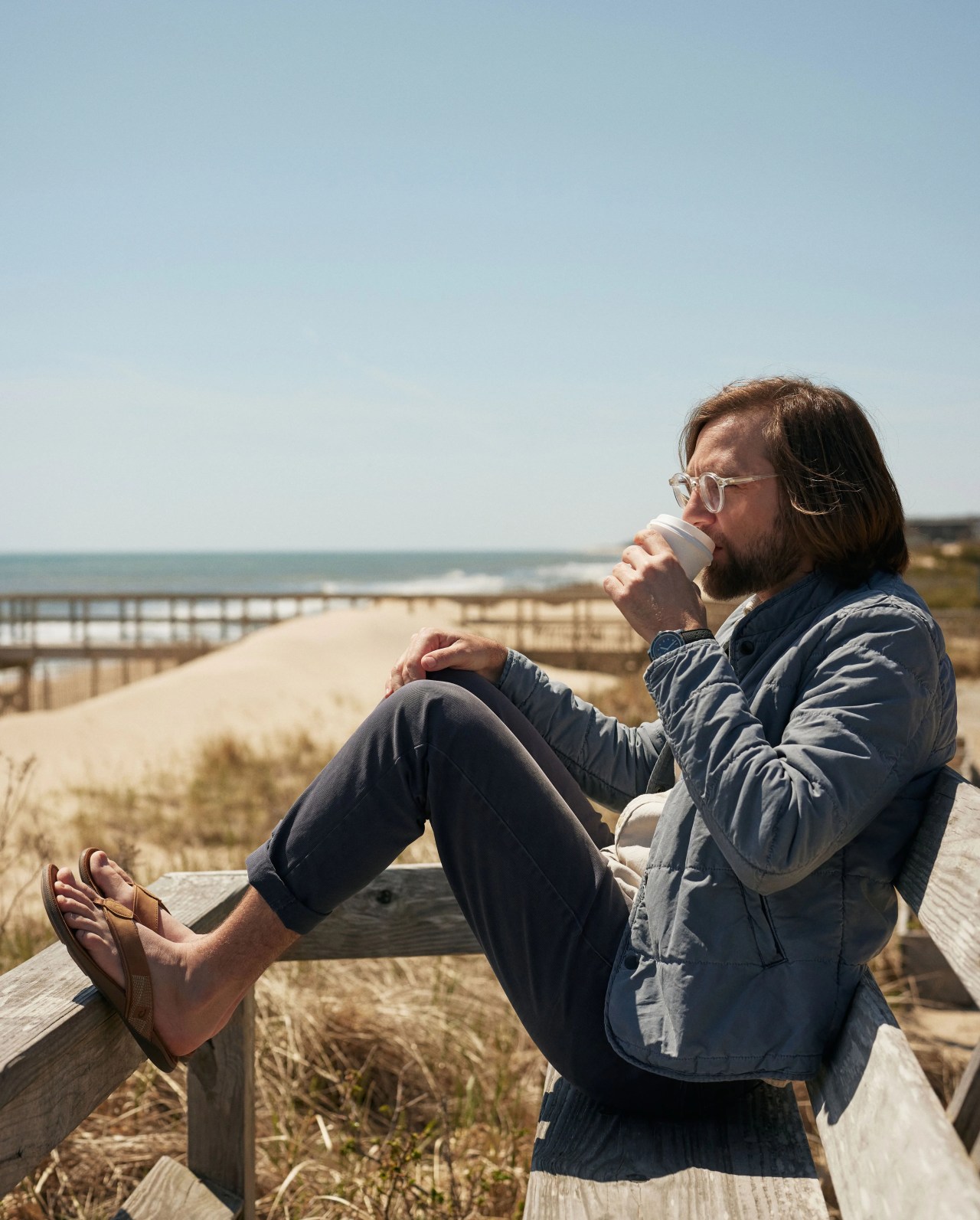mikey detemple sitting down at the beach while drinking some coffee wearing olukai tuahine sandals