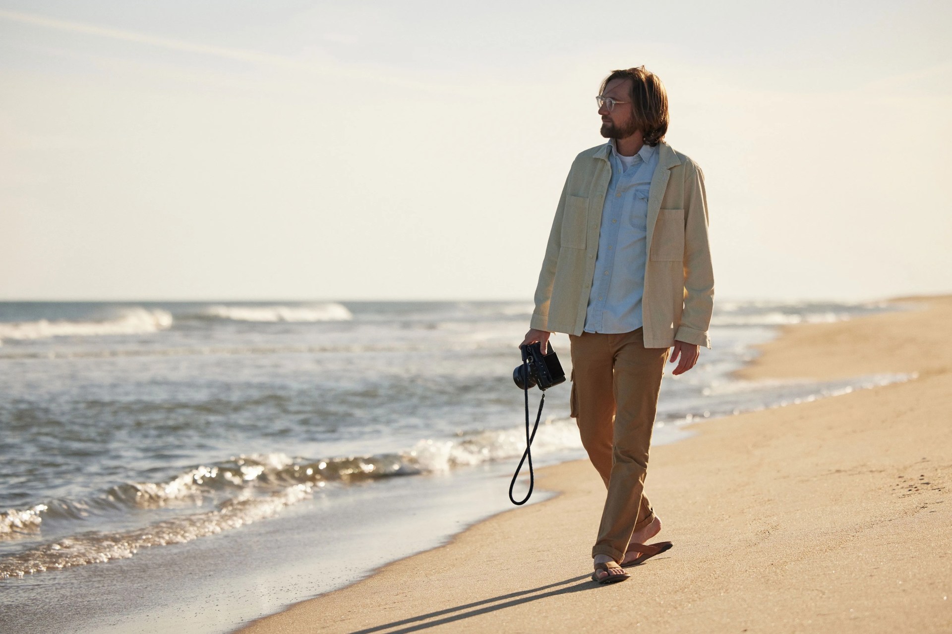 mikey detemple wearing olukai tuahine sandals walking along shoreline holding a camera in his right hand