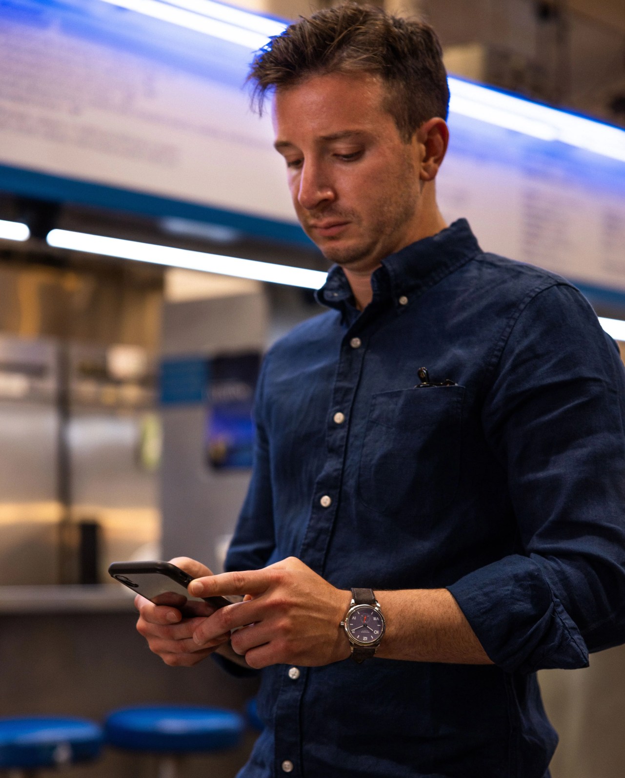 man standing by restaurant looking at phone wearing nomos watch and a blue dress shirt