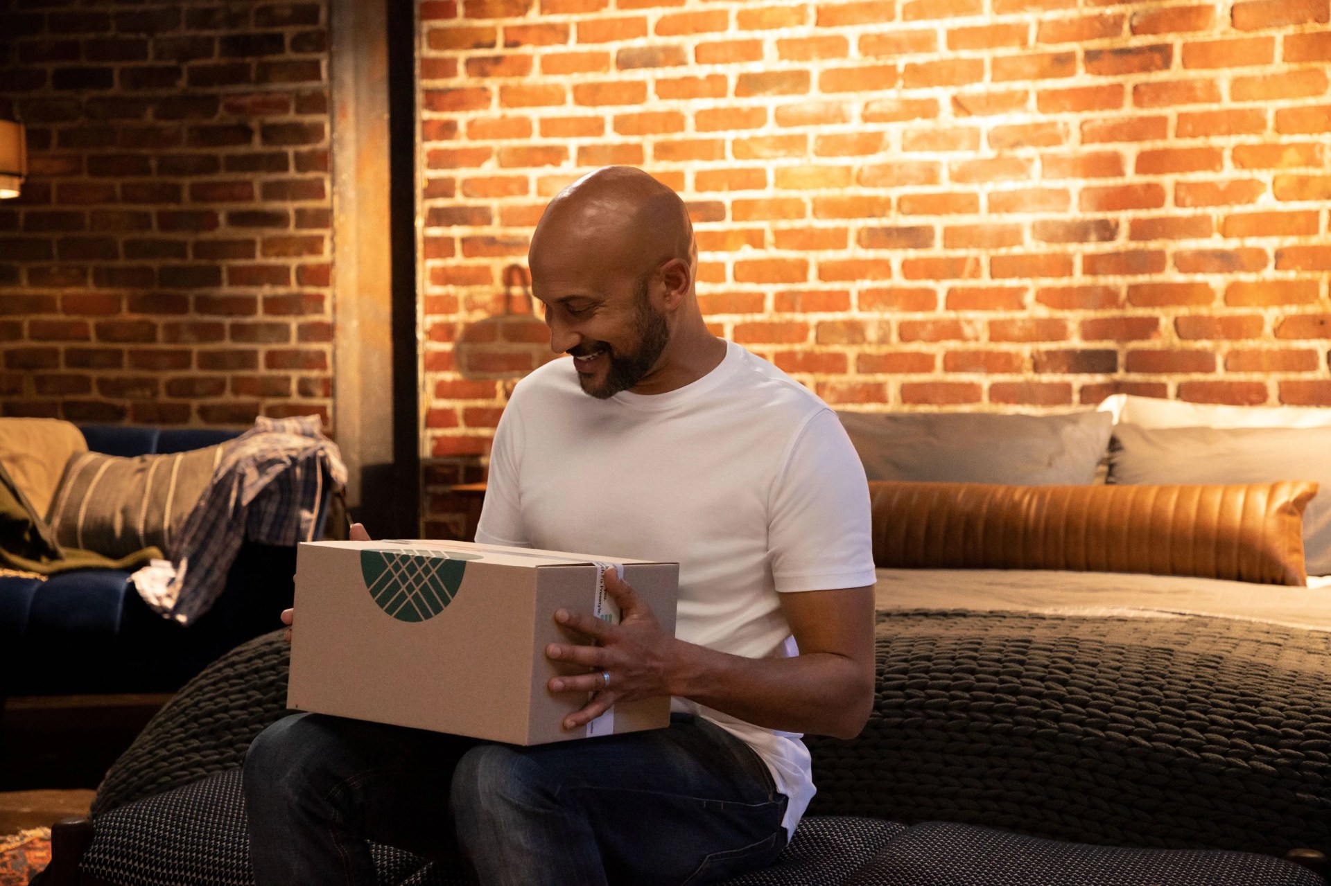 keegan michael key sitting down on edge of bed holding a stitch fix box in lap