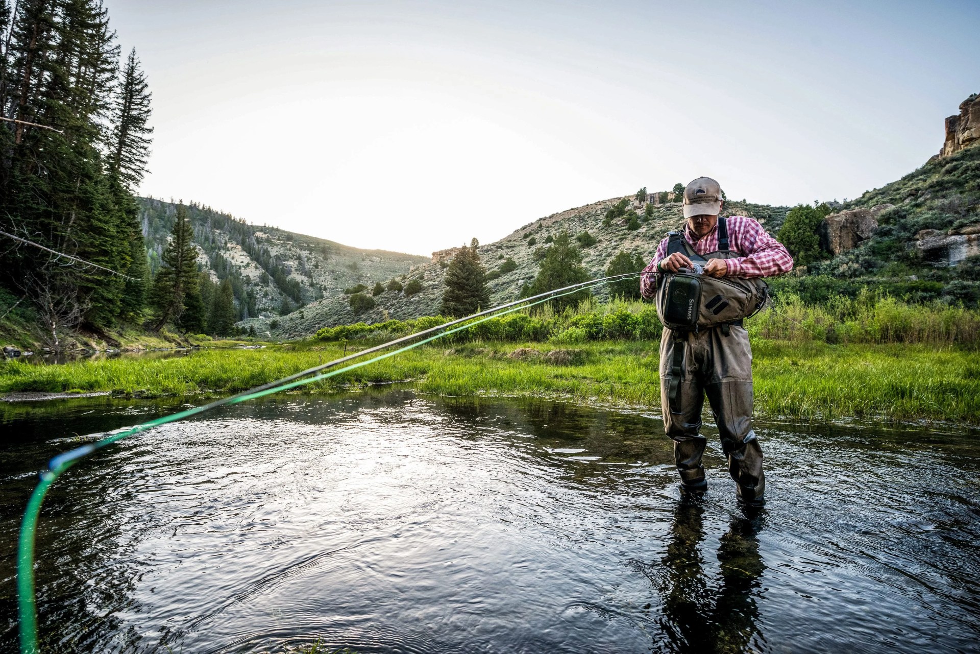 man standing in lake looking in bag while holding a fishing rod