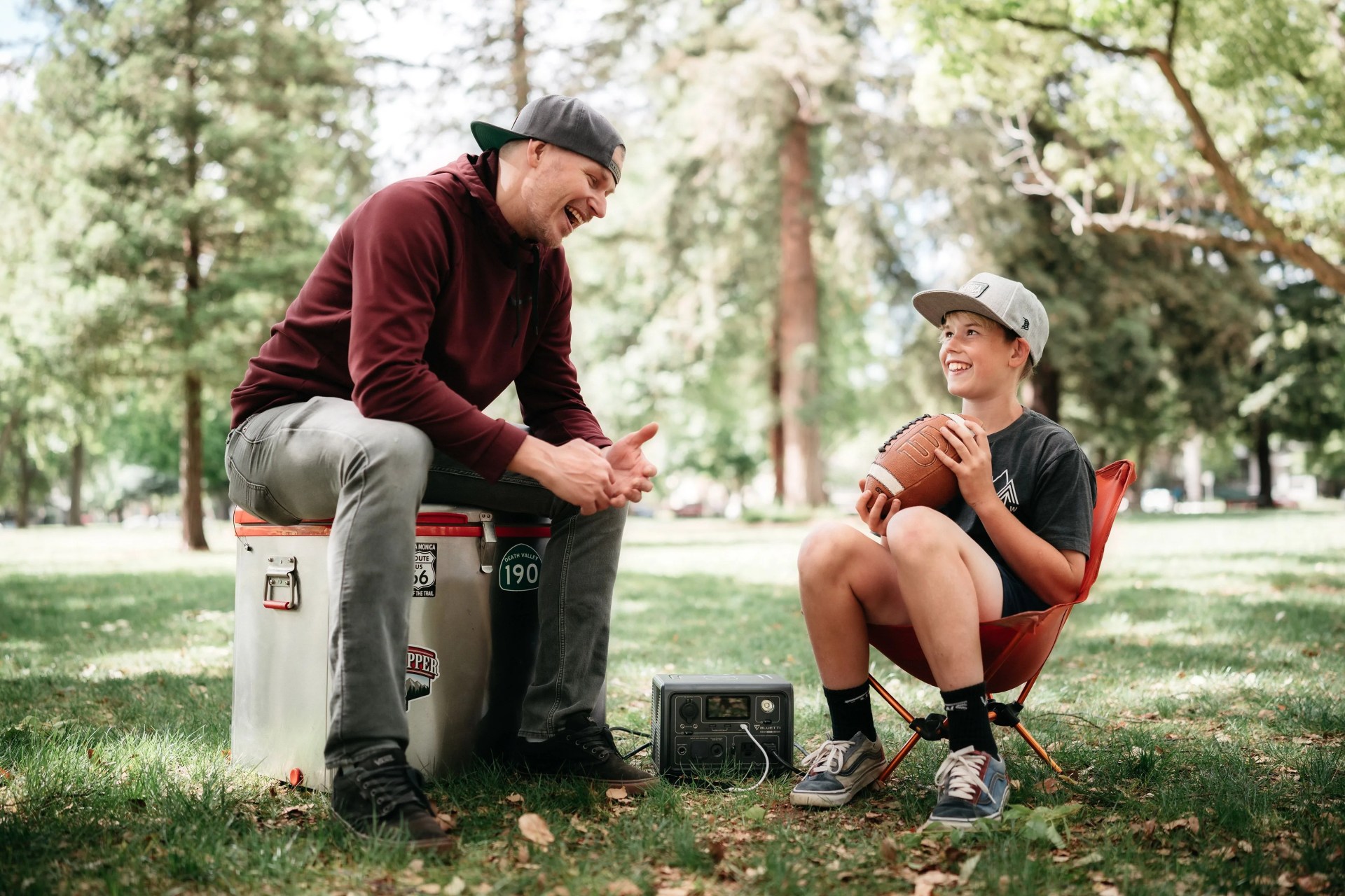 man sitting on cooler next to kid holding a football while sitting in a foldable chair in a field using bluetti charging station