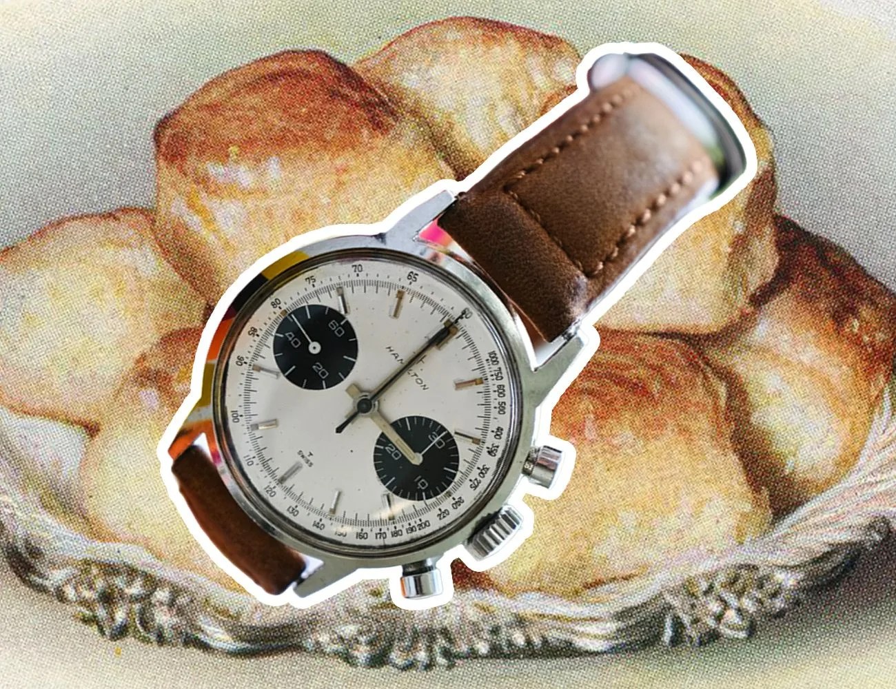 a silver hamilton intramatic panda chronograph watch with a brown leather strap overlayed on a background of biscuits