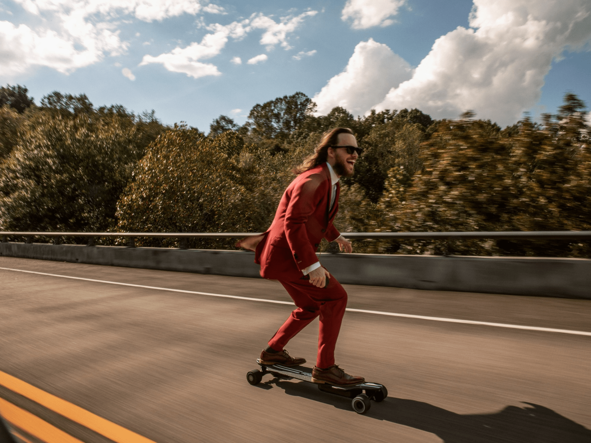 longhaired man skateboarding down street in sunglasses and red indochino suit