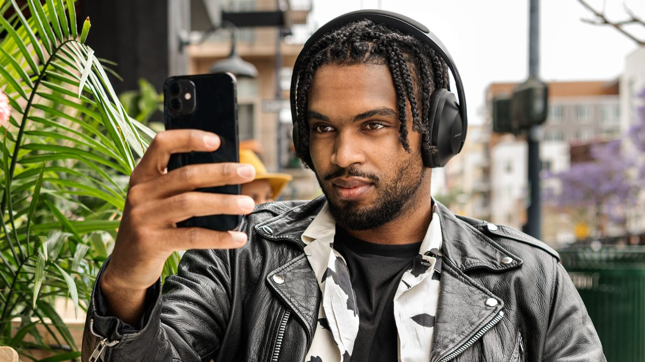 black man in leather jacket holding up cell phone wearing bose 700 headphones