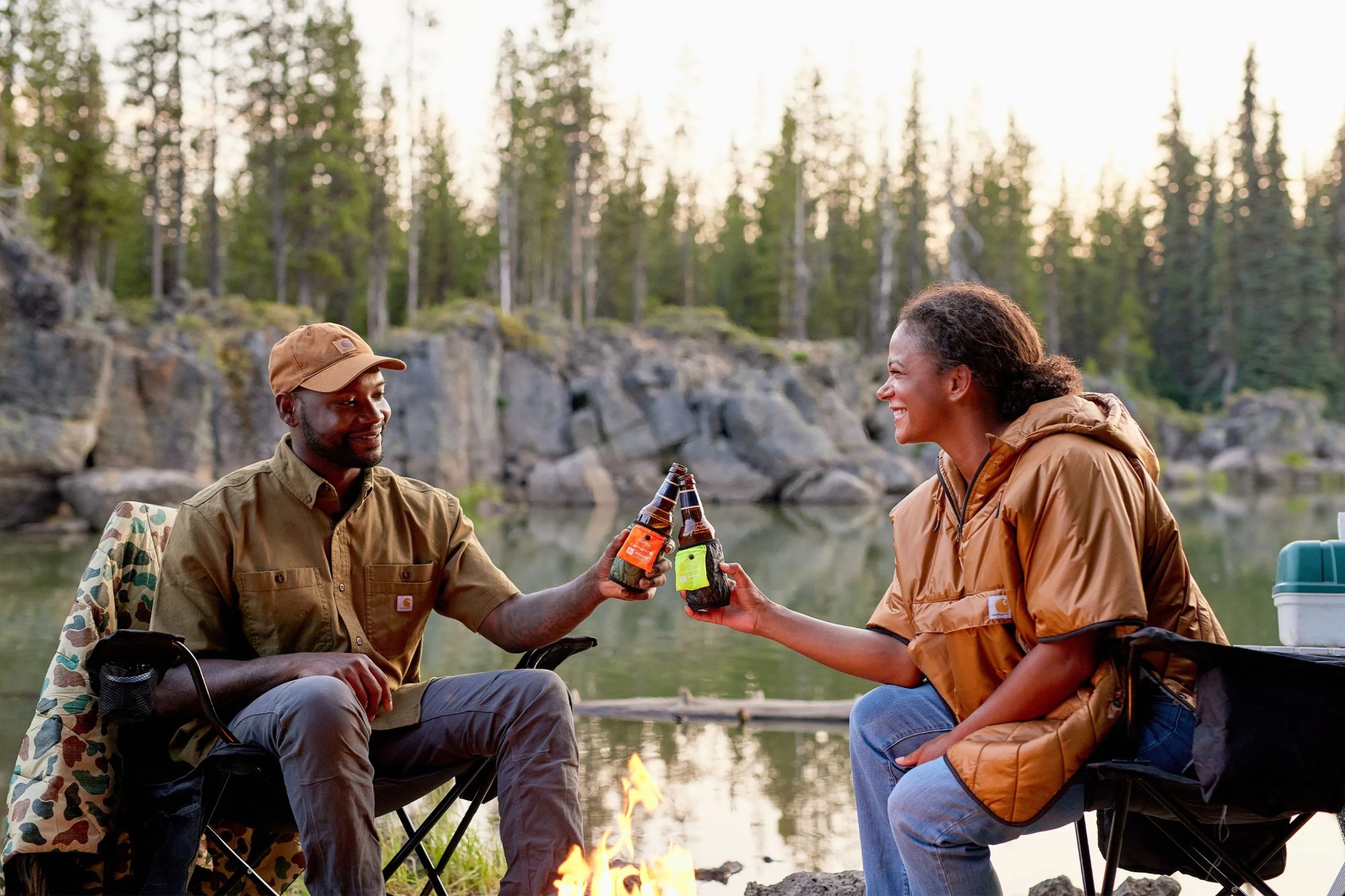 a couple sitting by a river cheersing beer bottles with carhartt x rumpl coozies