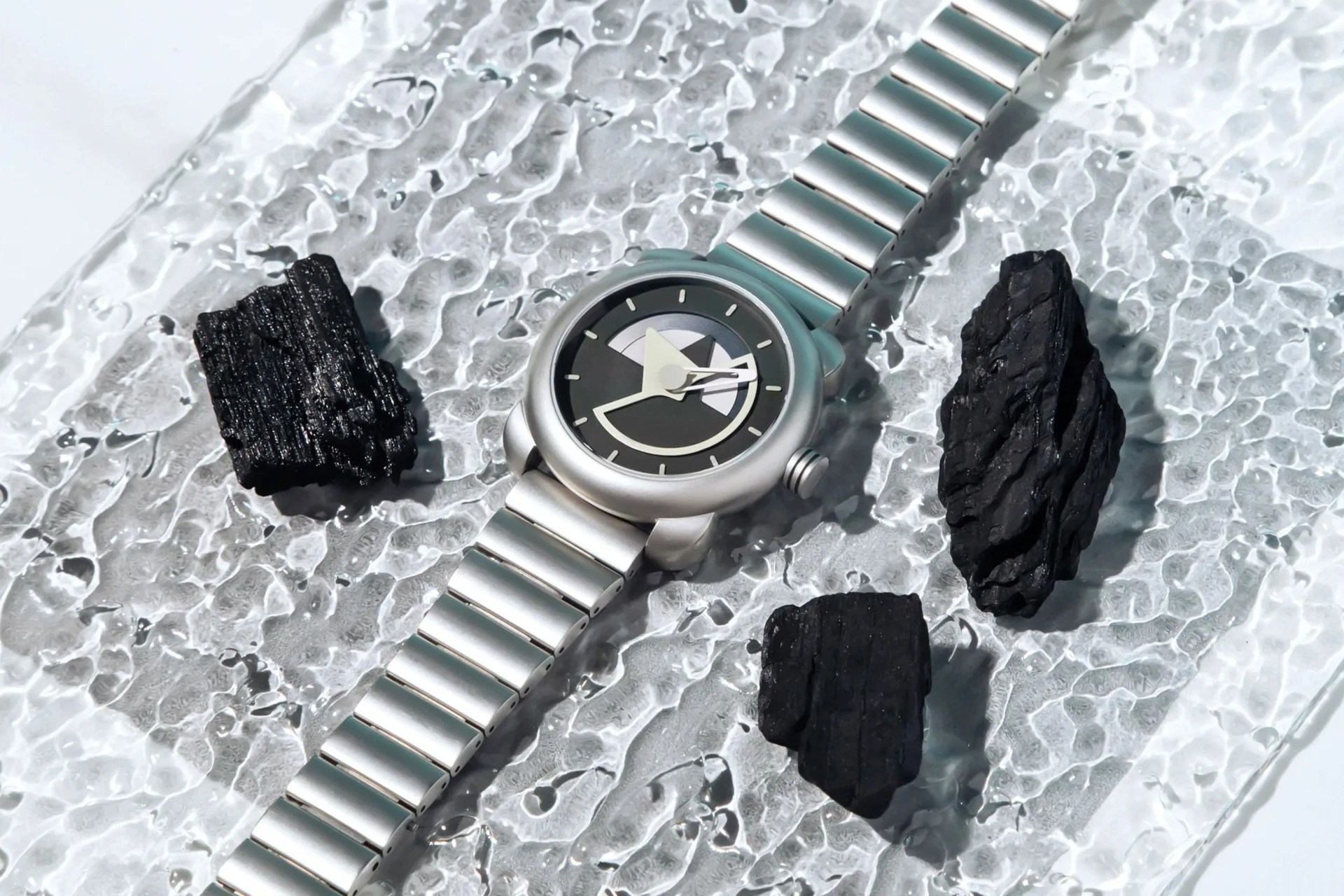 silver watch laying on glass with black lava stones around it