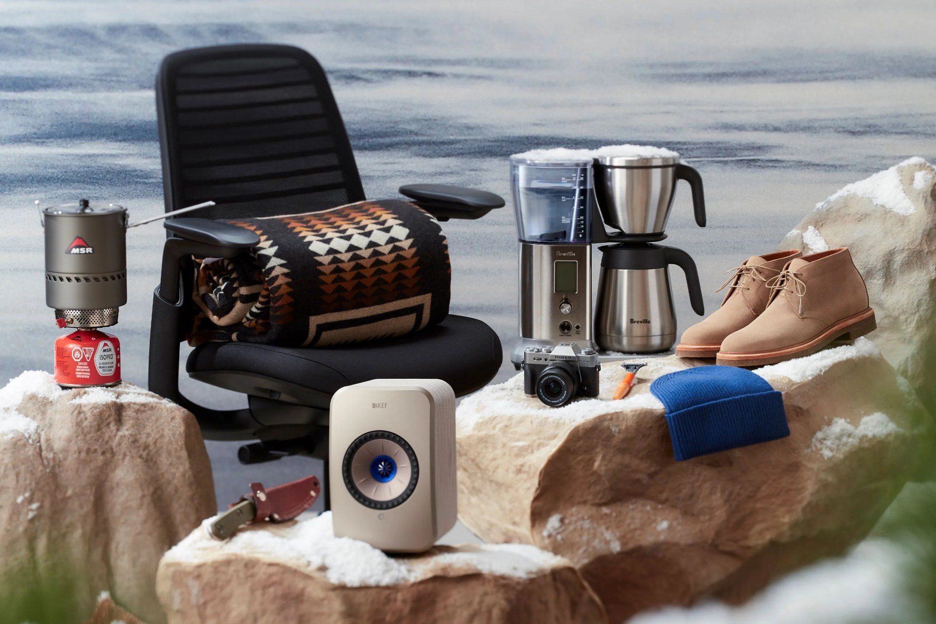 a portable water boiler, desk chair, blanket, coffee maker, shoes, camera, razor, blue beanie, knife, and speaker sitting on snowy rocks