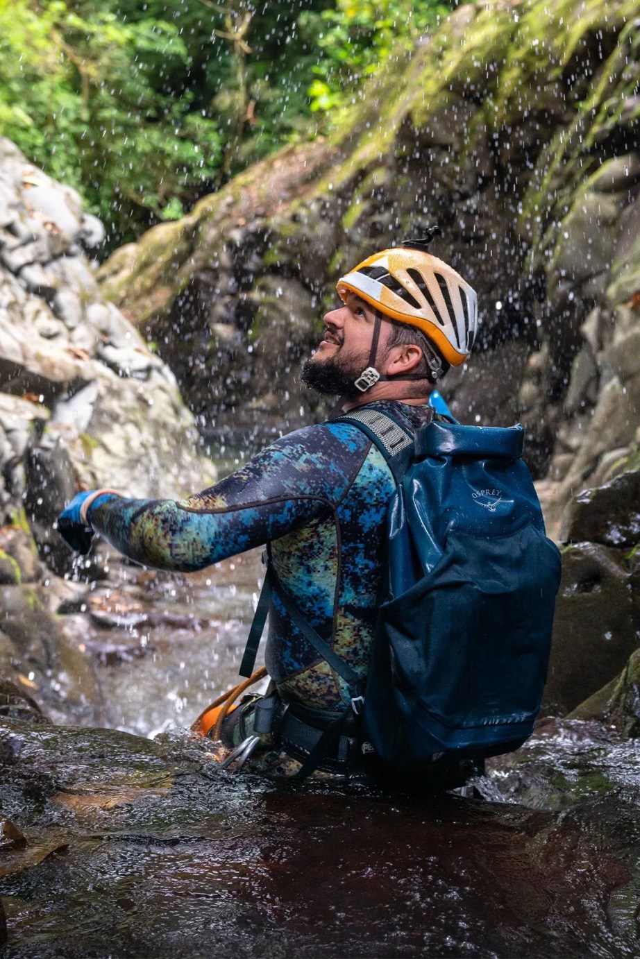 man walking through a river wearing a helmet and osprey backpack