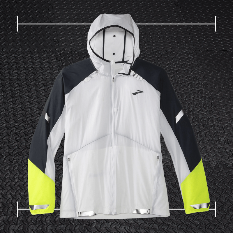 a white brooks running jacket with green and black accents laid over top a photo of black rubber gym flooring