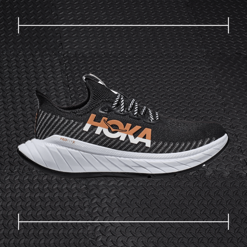 a black hoka shoe with a white sole and orange accents laid over top a photo of black rubber gym flooring