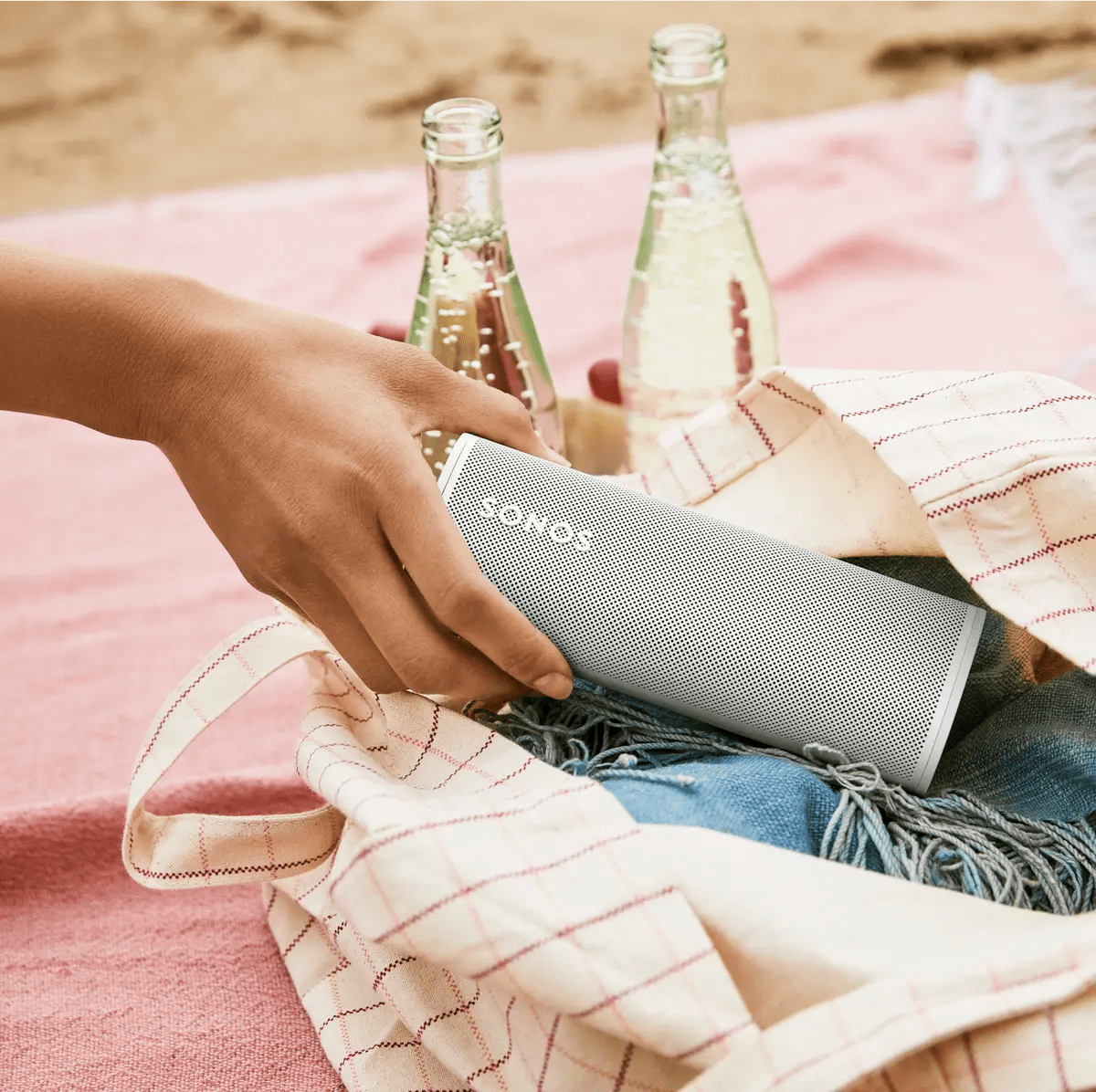 hand grabbing sonos roam from beach bag on towel in front of carbonated drinks with sand in the background