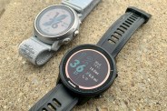 a silver coros watch laid on the ground next to a black garmin watch