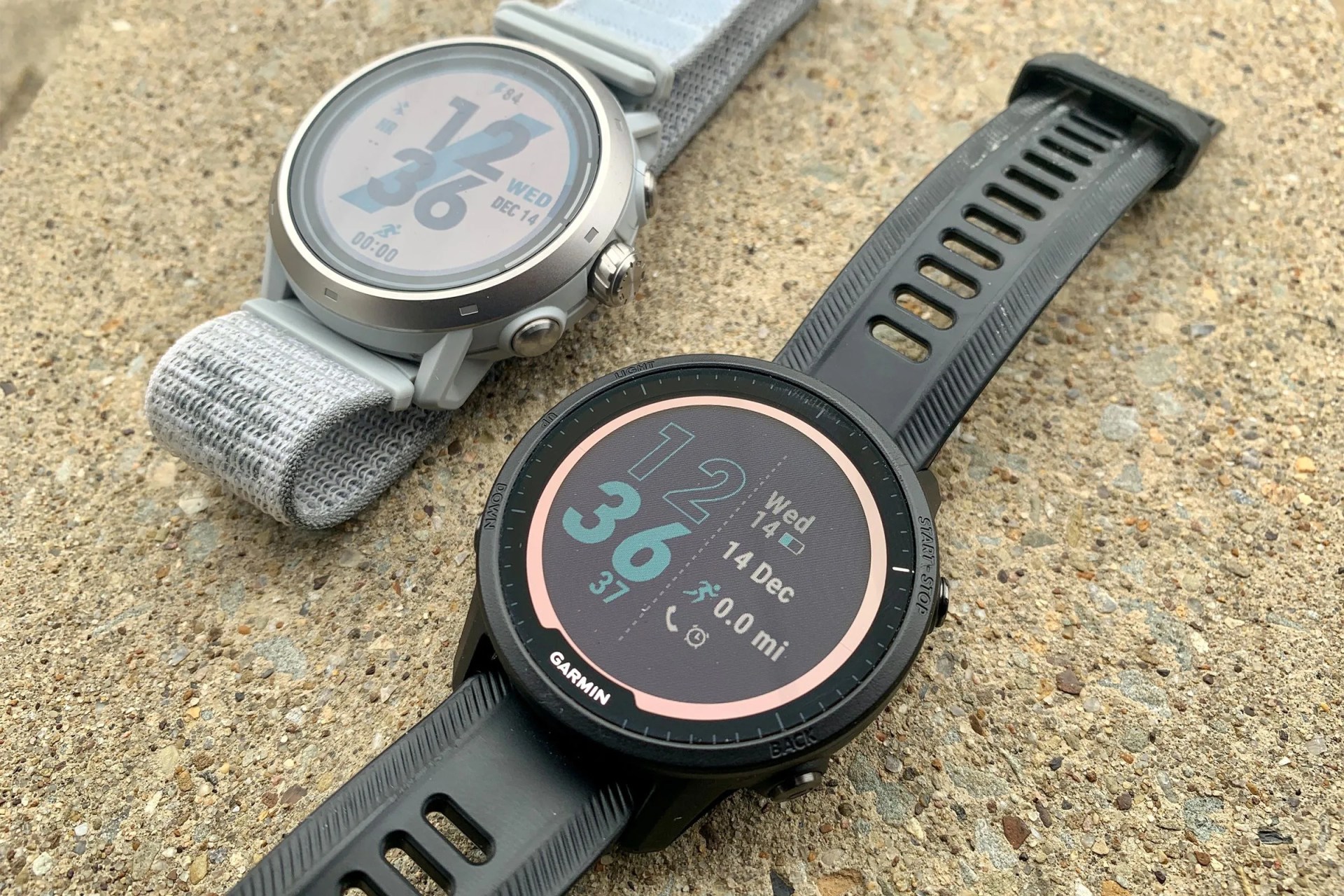 a silver coros watch laid on the ground next to a black garmin watch