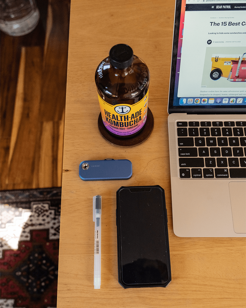 a close up of hayleys desk with a muji pen, phone, knife and kombucha laid out on a brown wood desk