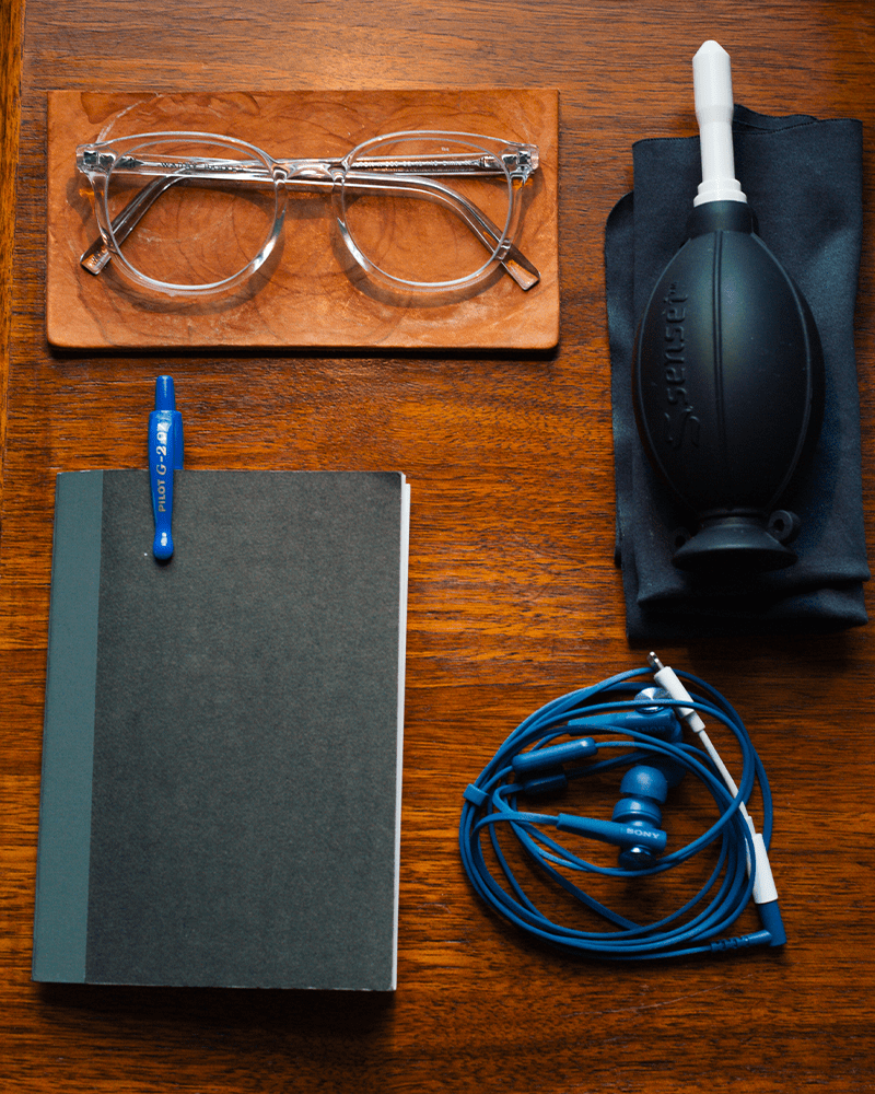 a desk with a black notebook, blue headphones, black cleaning bulb and transparent glasses on it