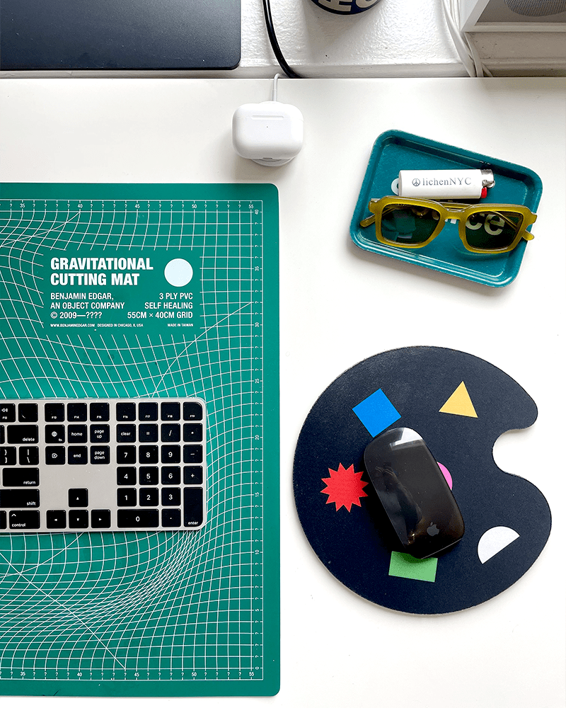 a close up of evans desk with a cutting mat, keyboard, mouse pad, mouse, sungless and airpods