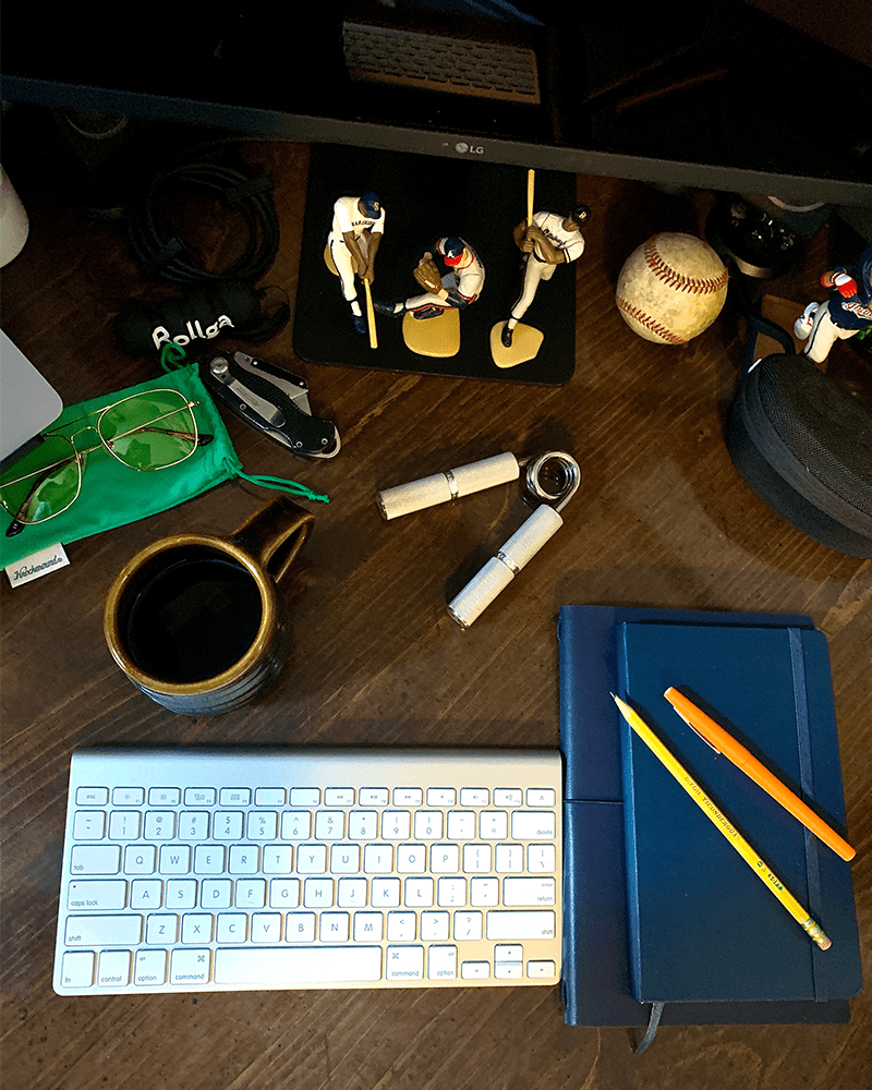 a close up of bens desk featuring a keyboard, coffee cup, pens, and notebook