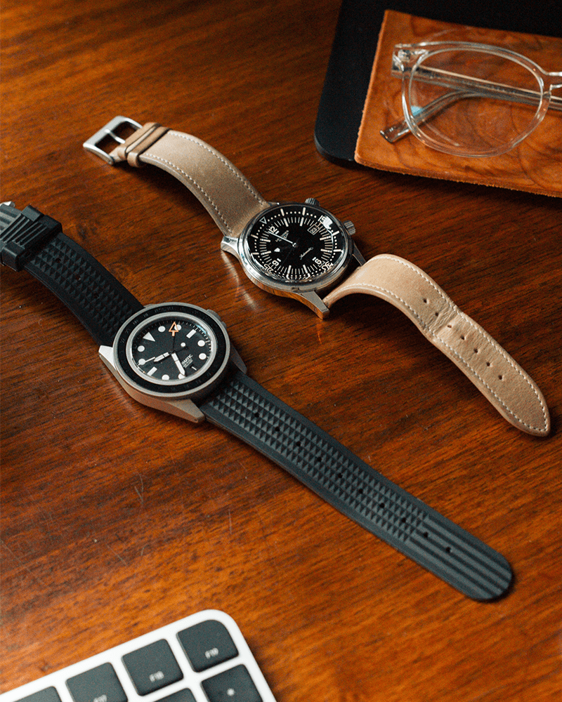a watch with a black strap laid next to a watch with a brown strap on a brown desk