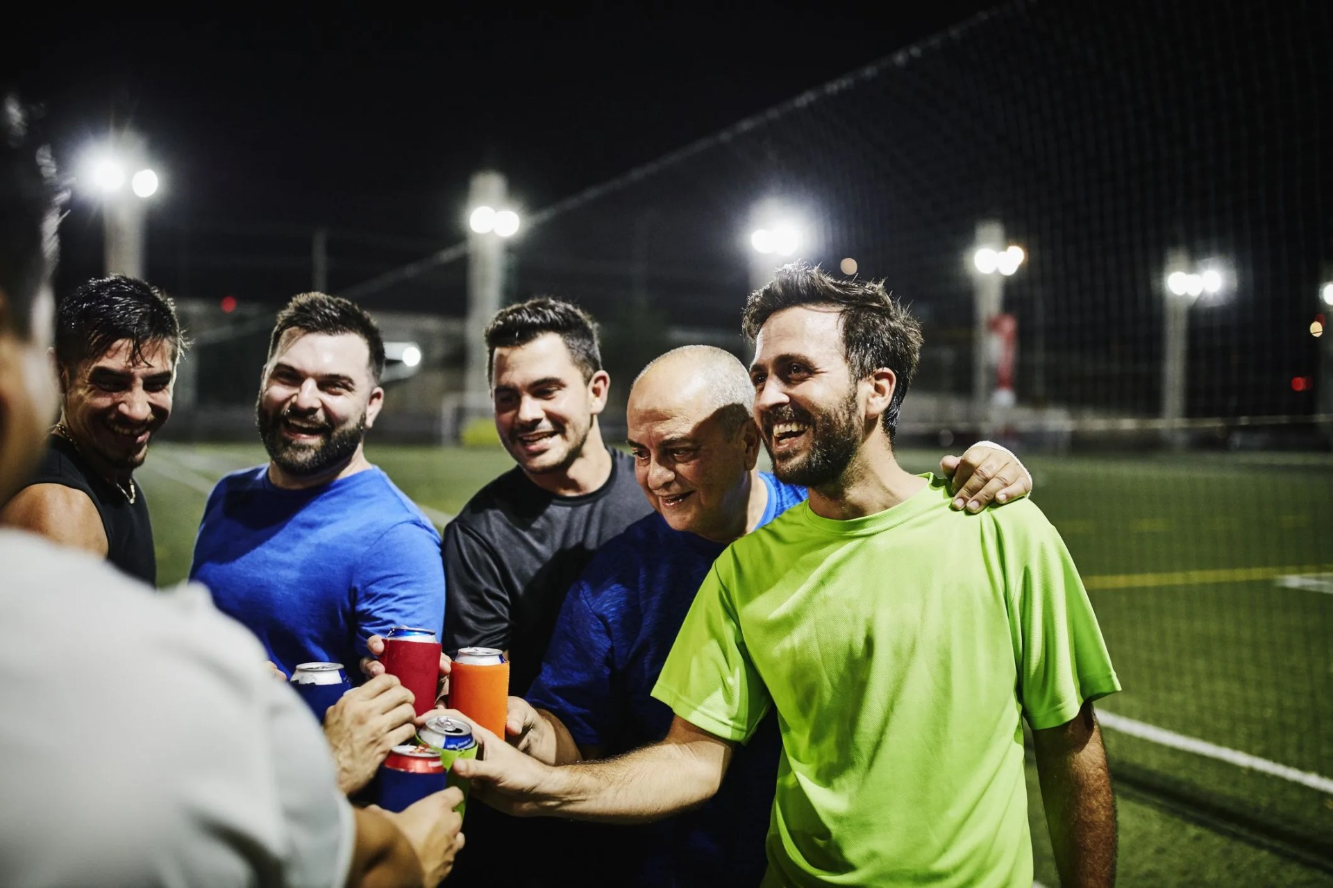 laughing male friends toasting with beers after nighttime soccer game