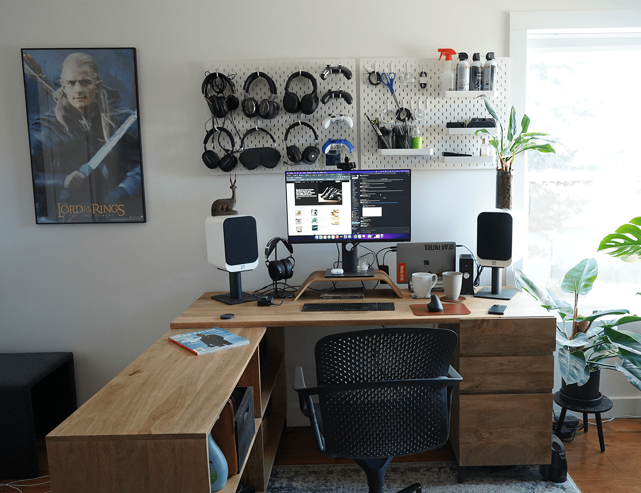 a light wood desk in front of a white wall with a lord of the rings poster and pegboard with multiple sets of headphones on it