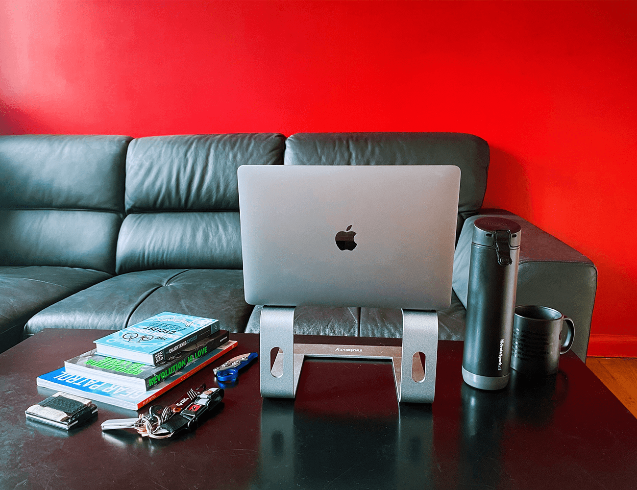 a laptop on a silver riser on a black desk in front of a black couch and red wall