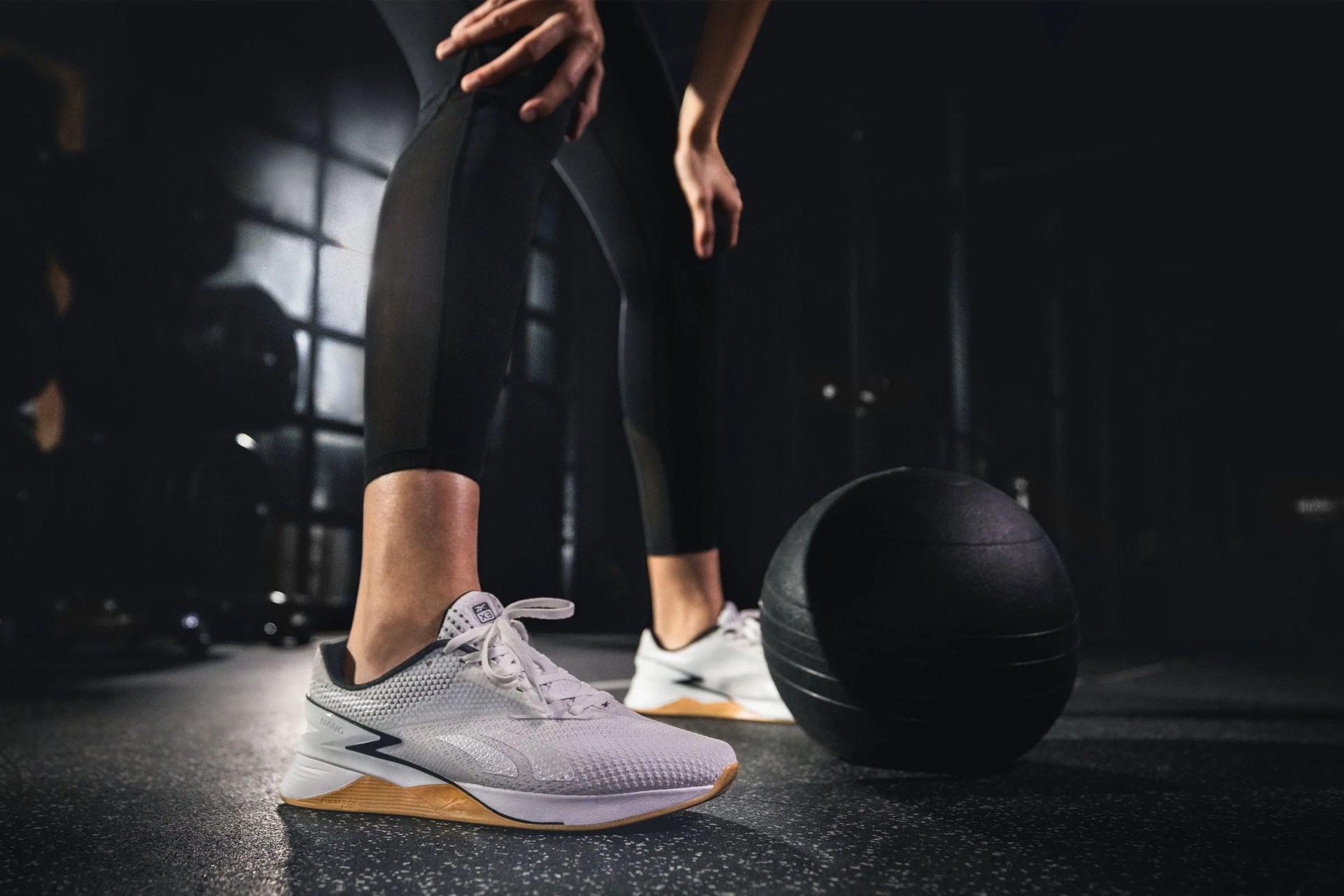 close up of a woman wearing white nano x3 shoes leaning over a weighted ball in a gym