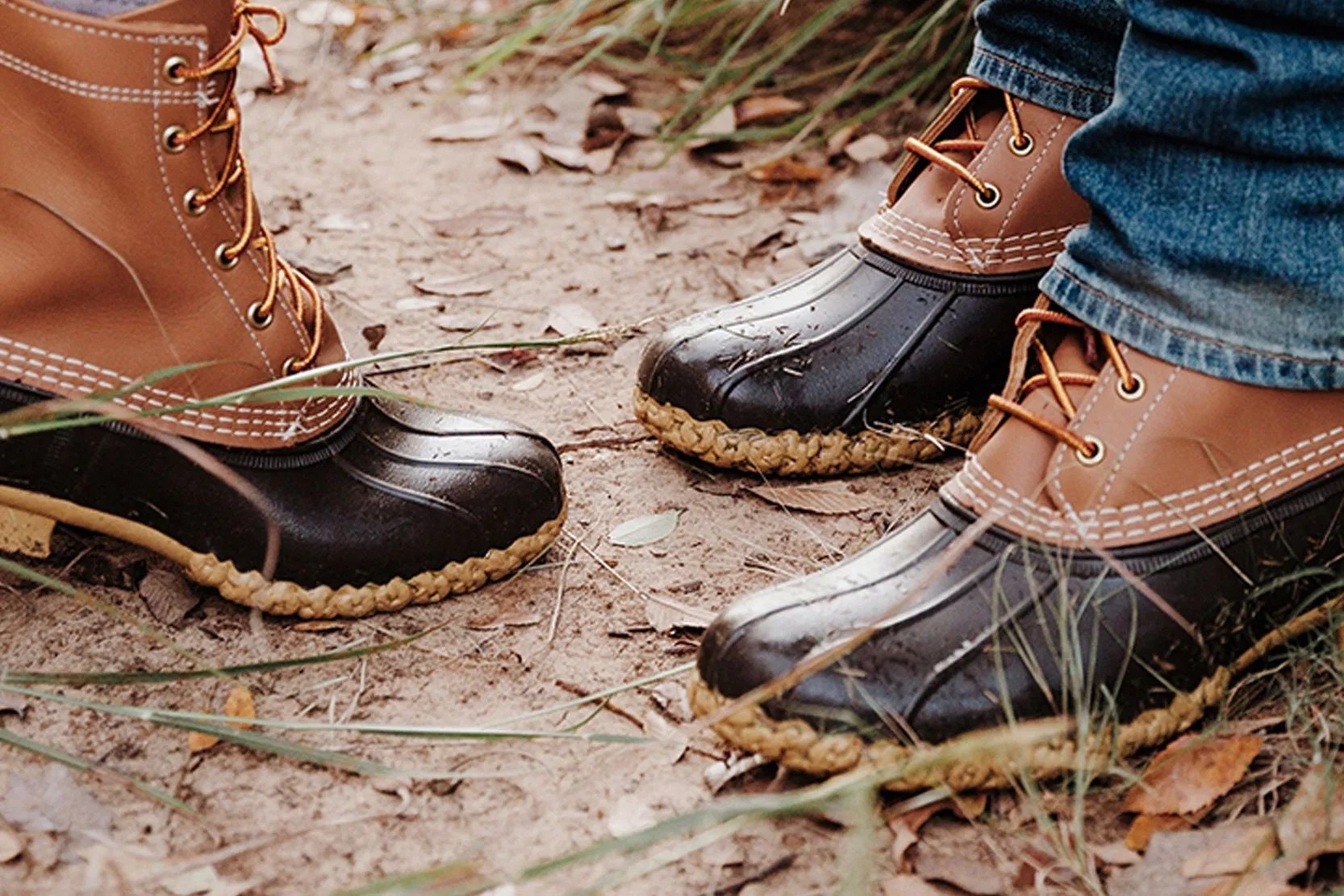 Two pairs of bean boots being worn by two individuals shown from the ankles down resting on a muddy ground.