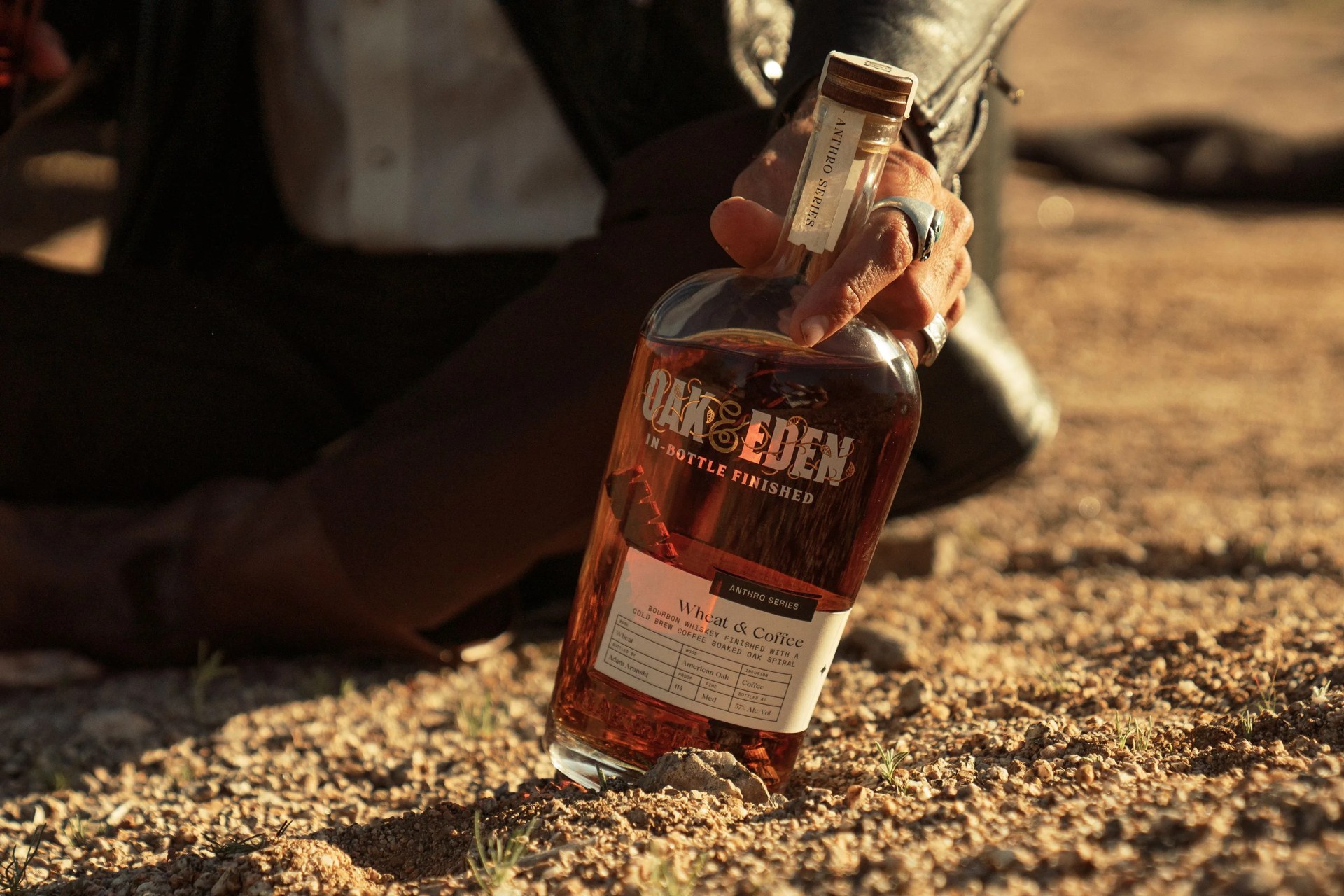 a man sitting in a desert landscape holding a glass bottle of oak and eden whiskey