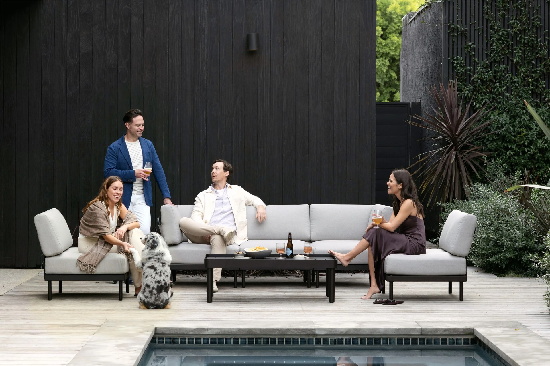 people sitting on a couch on a pool patio with drinks, snacks, and a dog