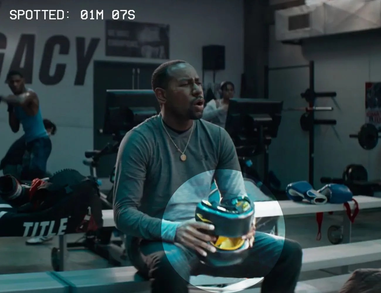 a man sitting on a bench in a gym holding an everlast elite headgear piece in black and gold