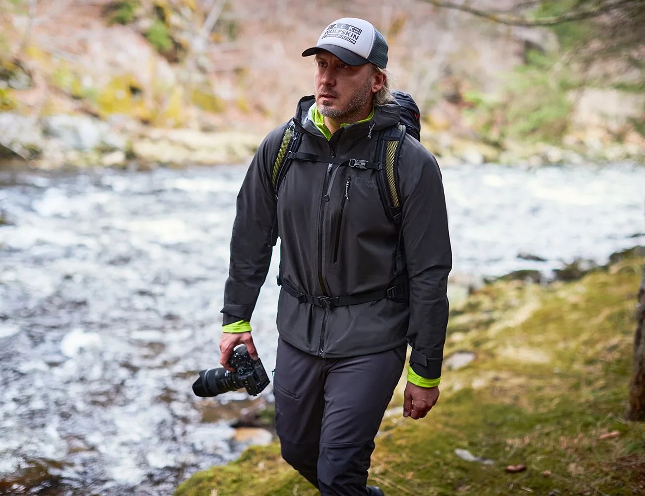 a man hiking wearing a black jack wolfskin jacket and white hat and holding a camera