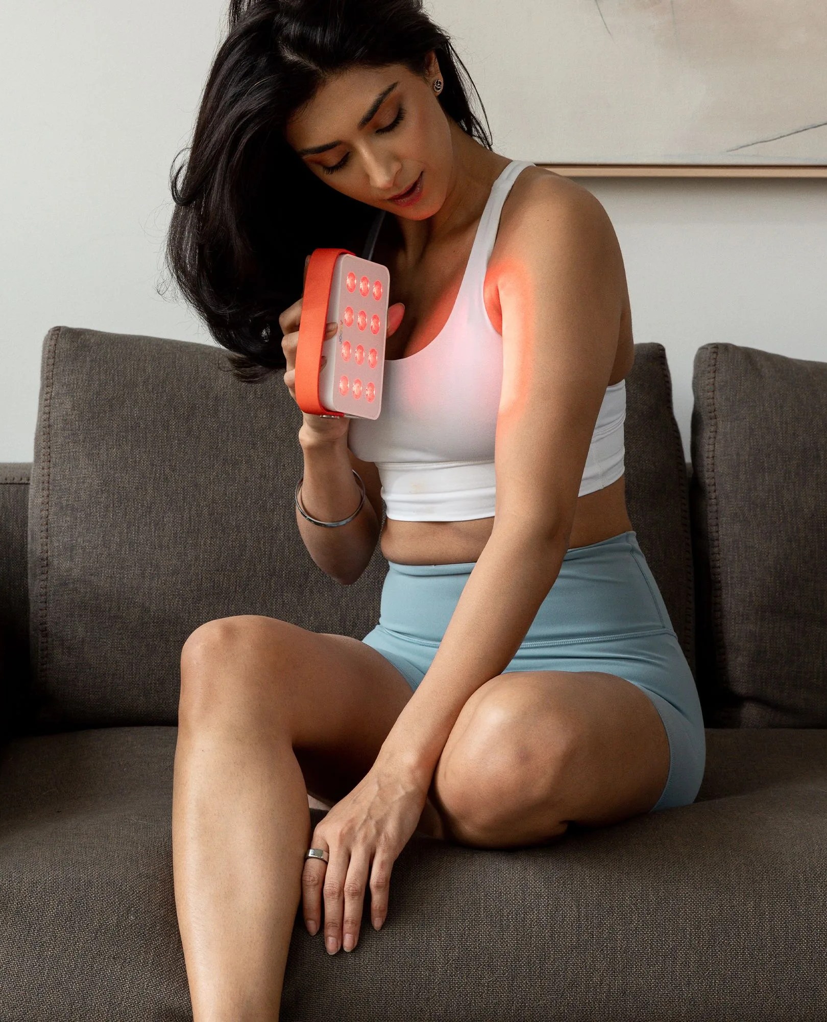 a woman sitting on a couch using a hand held red light therapy device on her arm