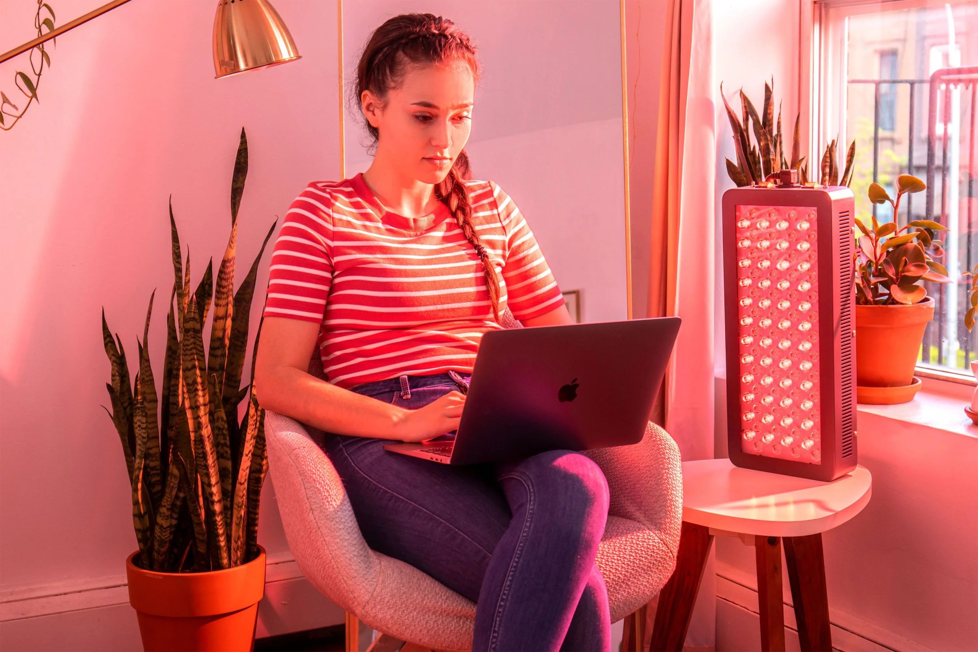 a woman working on her laptop with a red light therapy light on a table next to her