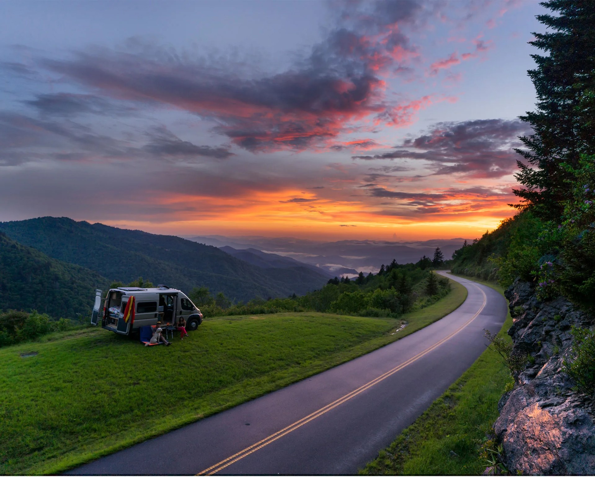 an rv parked on a grassy hill by the road during a sunset